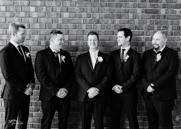 Groom and groomsmen by a brick wall, black and white